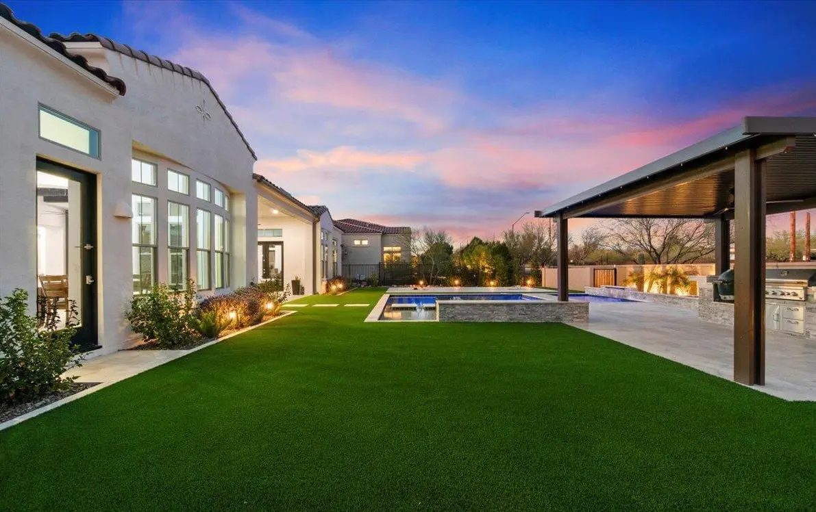 Modern backyard at sunset in Florence AZ with artificial grass, a rectangular swimming pool, and a covered outdoor kitchen area. The house features large windows and landscaped garden beds along the walkway.