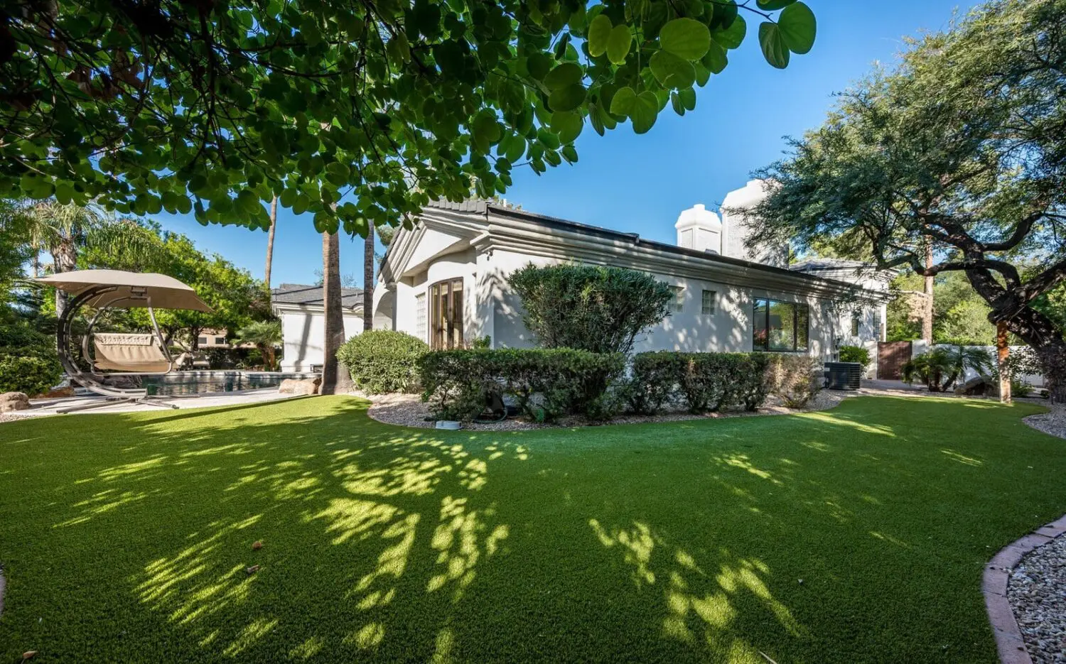 A backyard with Queen Creek artificial turf, leafy trees providing shade, neatly trimmed bushes along the side of a modern house, and a poolside daybed under a canopy in the background on a sunny day.