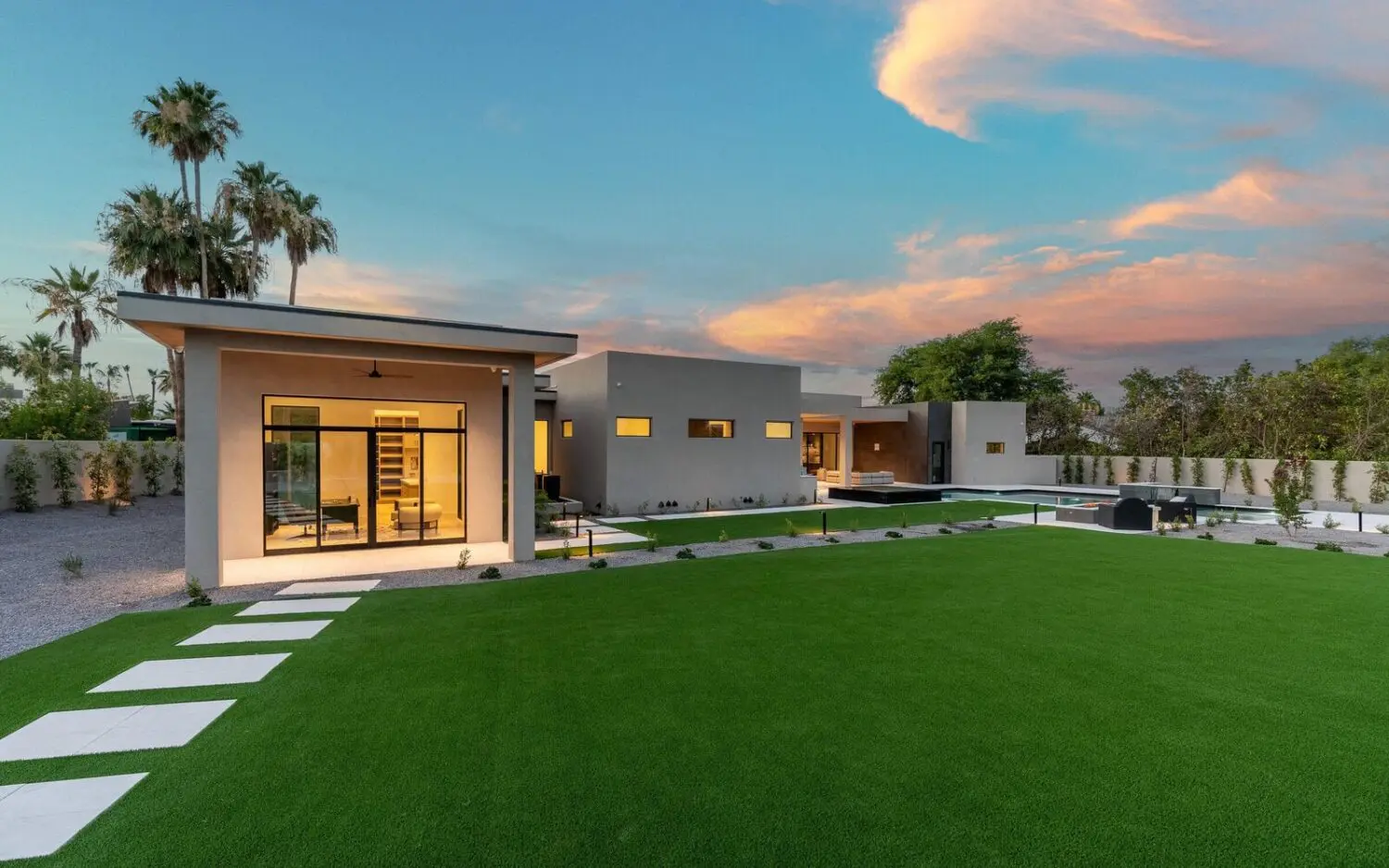 Modern single-story house with large windows, flat roof, and clean lines, set in a spacious Chandler AZ yard featuring pet turf and a stone path among palm trees, all under a colorful sunset sky.
