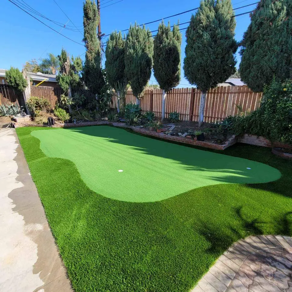 A backyard putting green with bright synthetic turf, surrounded by lush green grass and tall trees, sits beside a wooden fence under a clear blue sky—perfect for putting greens enthusiasts in Gilbert, AZ.
