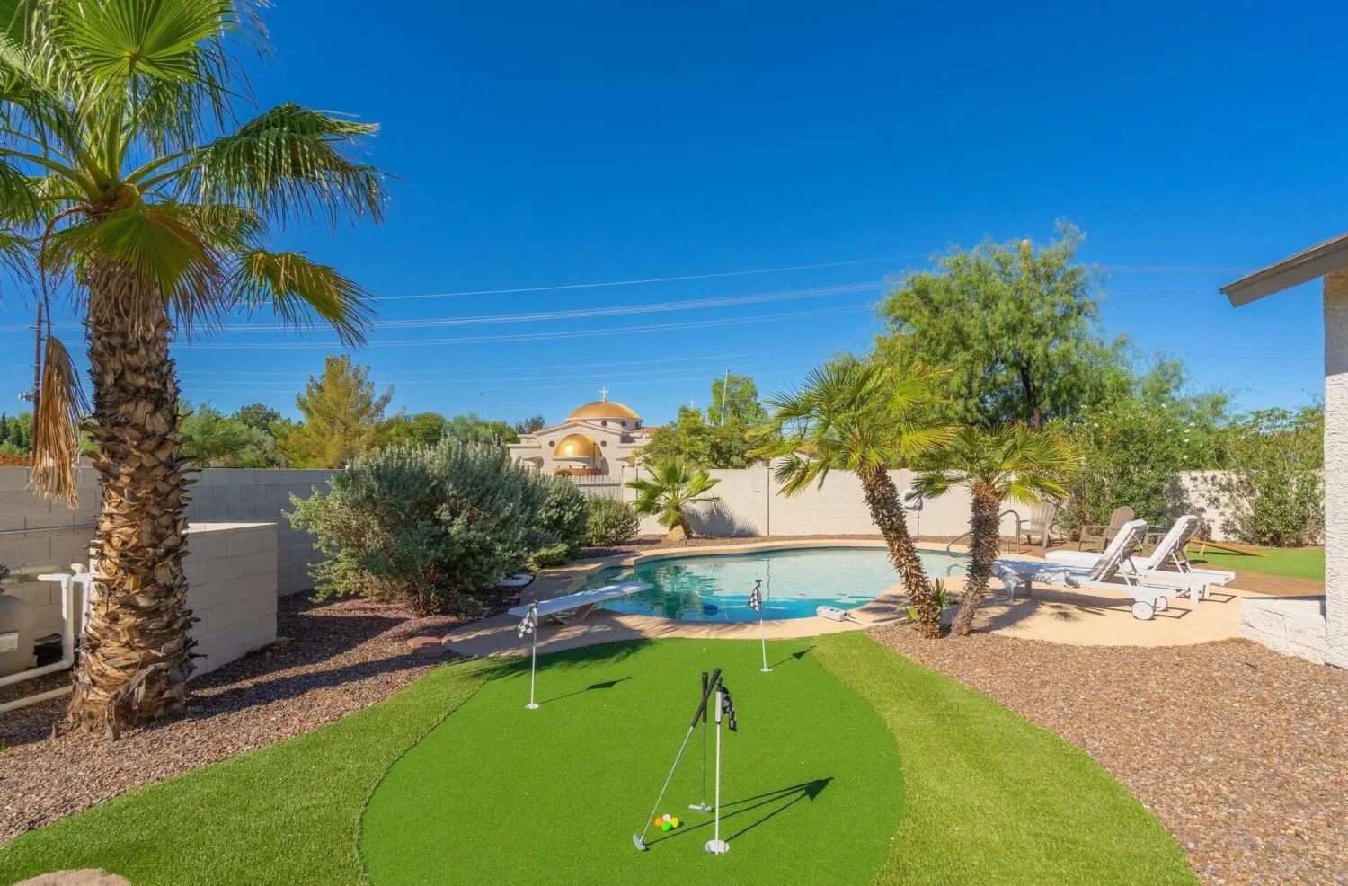 A backyard featuring a small putting green installed by expert artificial turf installers, palm trees, desert landscaping, and a swimming pool with lounge chairs under a clear blue sky.