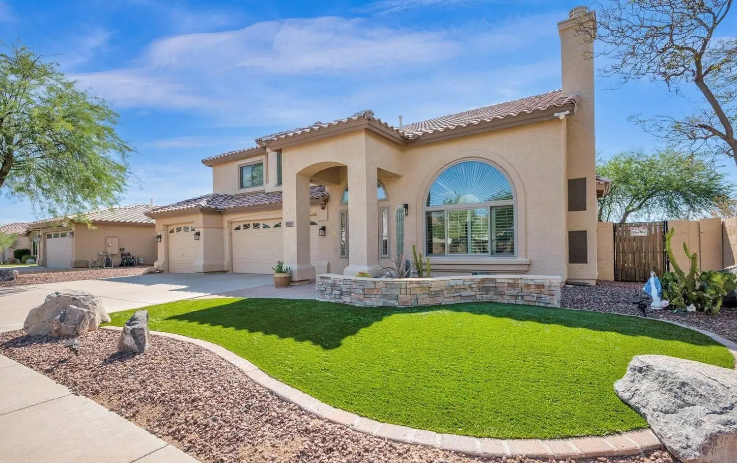 A modern beige stucco house in Apache Junction with a three-car garage, arched entryway, large decorative window, and a neatly landscaped front yard featuring synthetic turf installation and desert plants under a bright blue sky.