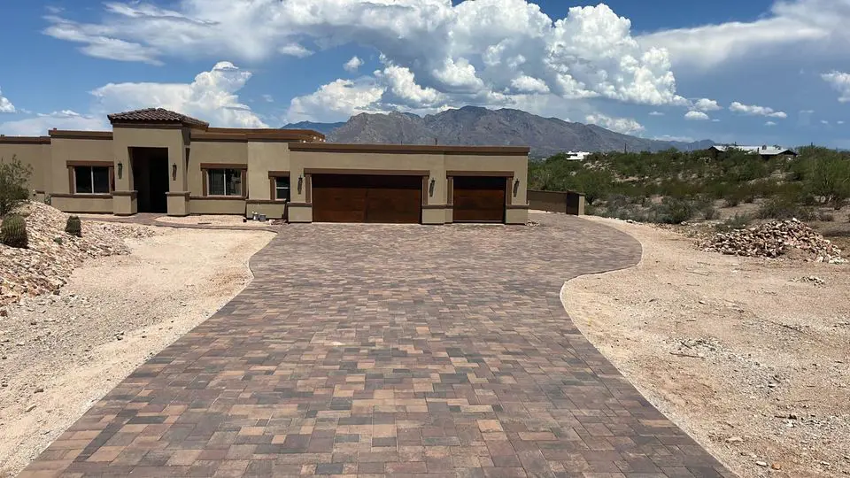 A modern, single-story house with a large, paved stone driveway sits amid a desert landscape enhanced by lush artificial grass. Mountains and a partly cloudy sky are visible in the background.