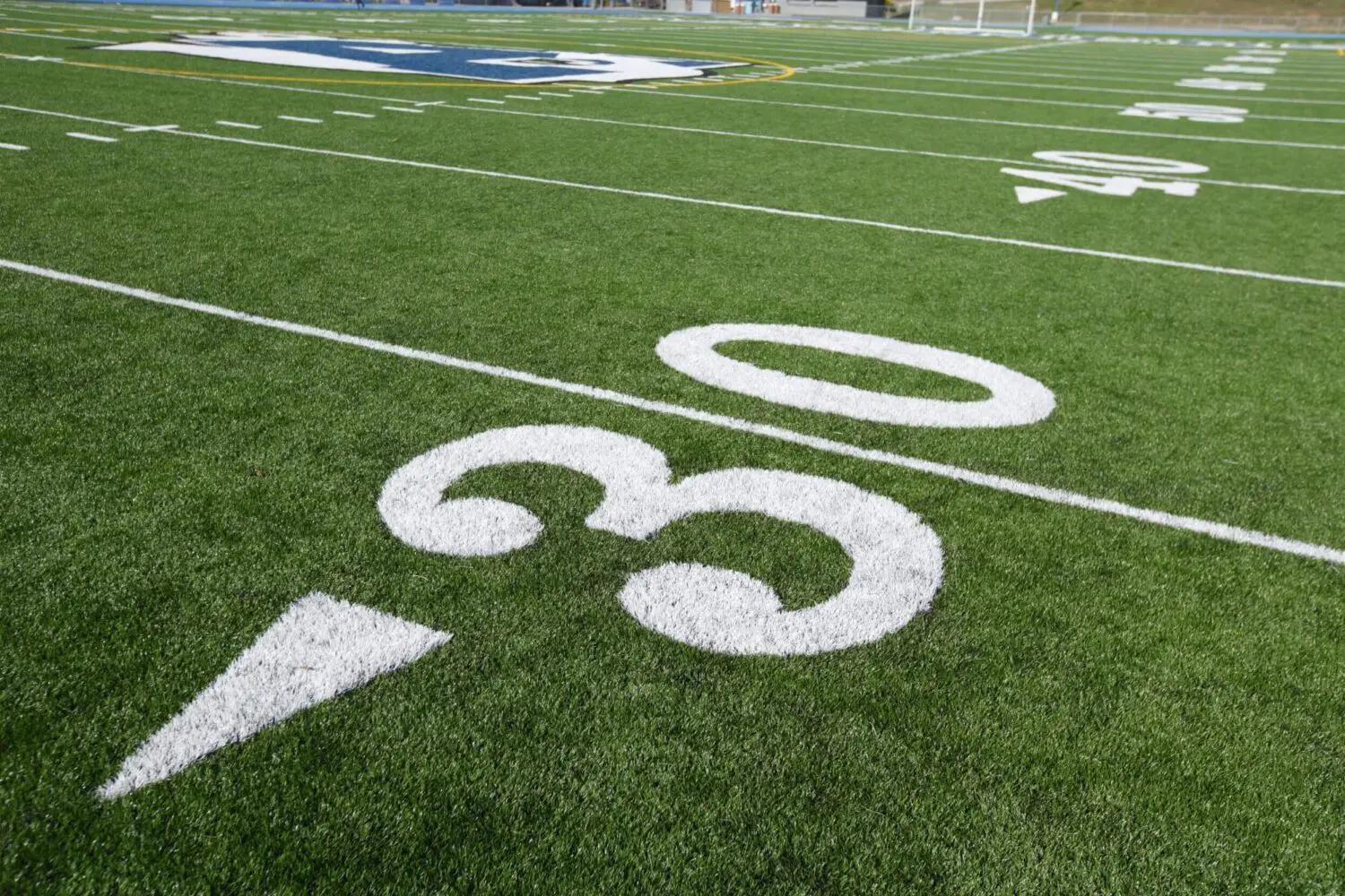 Close-up view of the 30-yard line on an American football field with bright white markings on green artificial grass installation. The field's logo is visible in the background, showcasing quality turf like that used for pet turf in Chandler AZ.