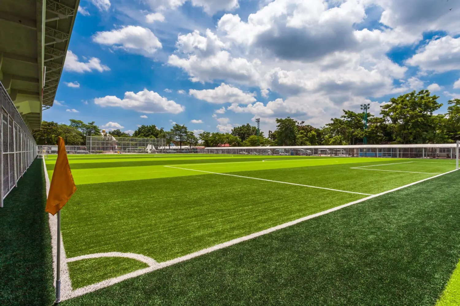 A bright, sunny day at an empty outdoor soccer field in Florence AZ with lush green artificial grass, crisp white boundary lines, a corner flag, and a clear blue sky dotted with scattered clouds overhead.
