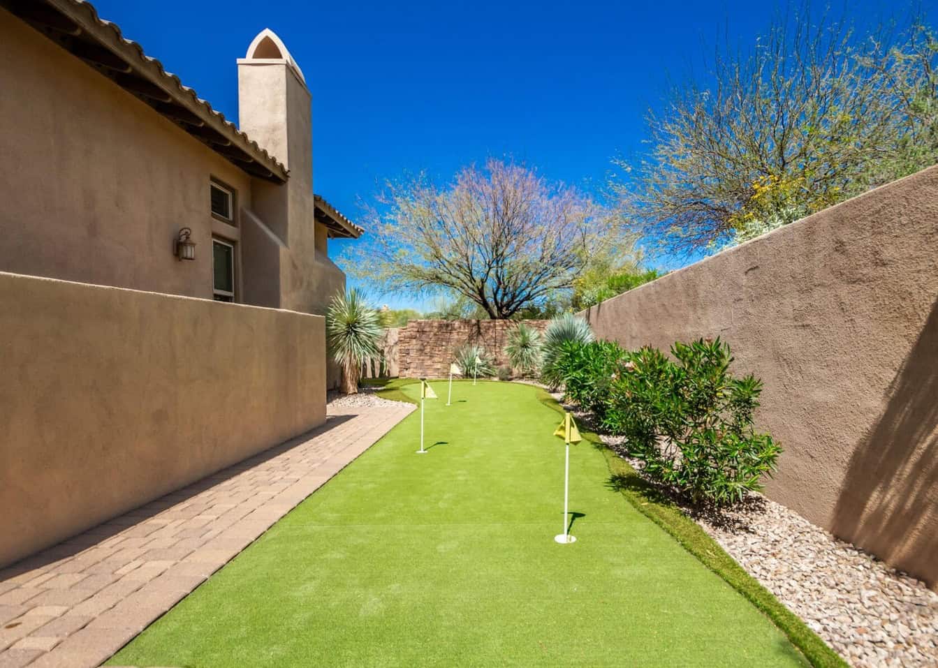 A narrow backyard in Apache Junction features a putting green with three holes, surrounded by desert plants, tan stucco walls, and a paved walkway—expertly finished with professional synthetic turf installation under a bright blue sky.