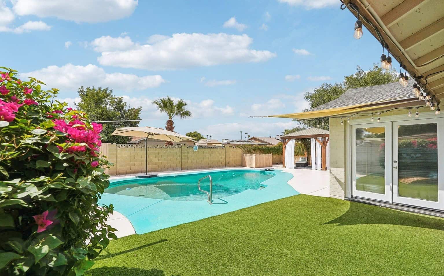 Backyard in Gilbert AZ with a swimming pool, synthetic turf, patio string lights, a gazebo, and blooming pink bougainvillea under a blue sky with scattered clouds.