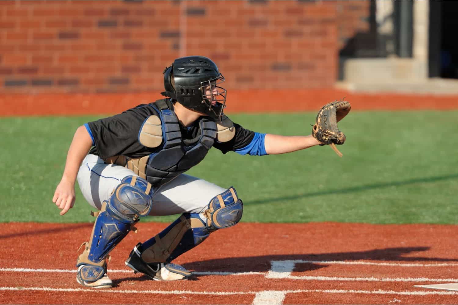A baseball catcher in full gear crouches behind home plate on a field of artificial grass Gilbert AZ, wearing a helmet, chest protector, and shin guards, with a glove extended to catch the ball.